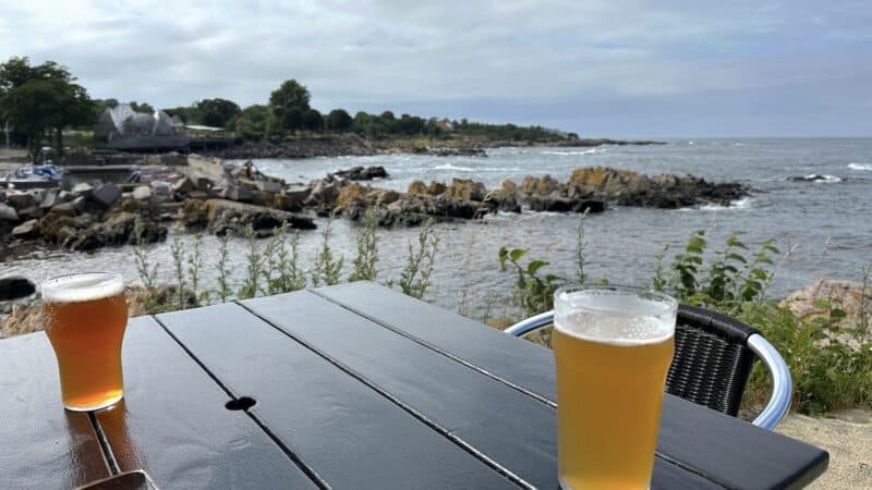 Zwei Gläser Bier auf einem Tisch in der Allinge Røgeri mit Ausblick aufs Meer mit Felsen am kleinen Kampeløkke Havn, Allinge