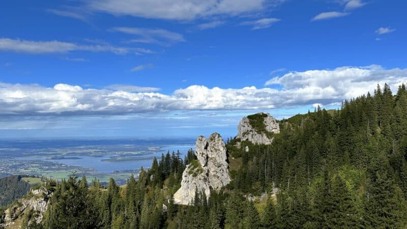 Titelbild: Ausblick von oben auf den Chiemsee mit einem großen Felsen an der Seite, eingebettet von Wald