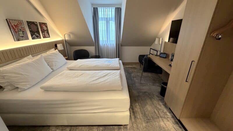 Dorint Hotelzimmer in gedämpften Brauntönen, weisss bezogenes Bett und ein großes Fenster gegenüber der Türe